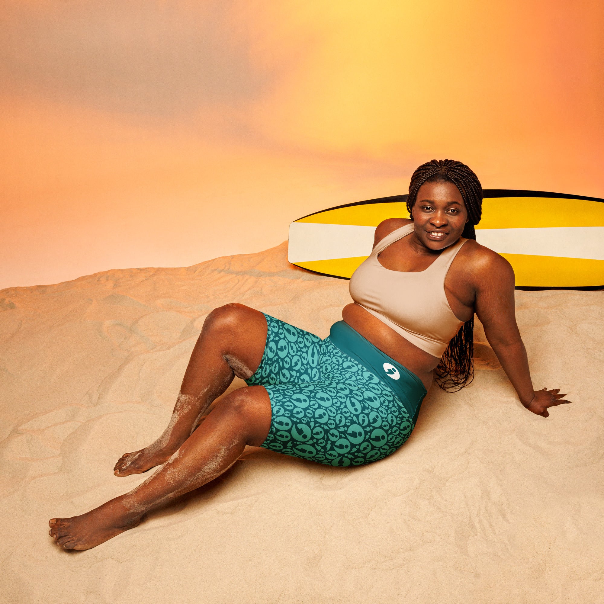 Model wearing teal patterned shorts sitting reclined with her hands propping her up with her legs slightly bent.