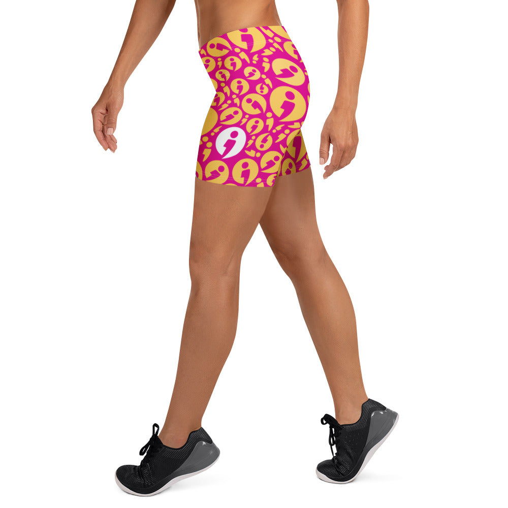 A person pictured from torso below wearing The Movement Collection's fitted shorts in the pink color variant featuring bright pink semi-colon avatars randomly placed over a yellow-orange background.
