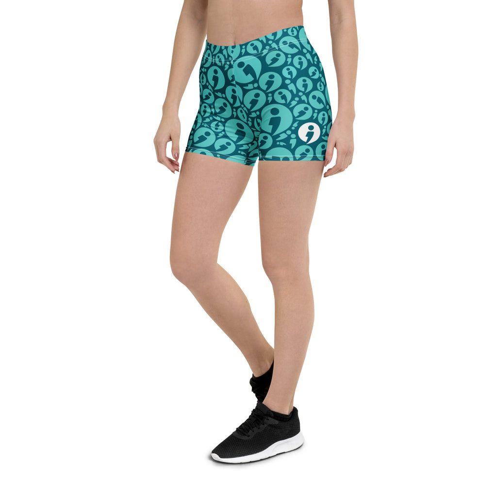 A person pictured from torso below wearing The Movement Collection's fitted shorts in the teal color variant featuring light blue semi-colon avatar randomly placed over a darker teal background.