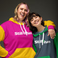 Two people wearing the Movement Collection unisex hoodies. Both feature the 'Still I Run' logo. The person on the left is wearing the pink color variant and the person on the right is wearing the gray color variant.